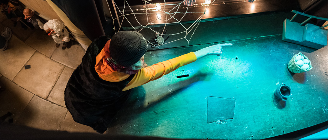 Hinter den Kulissen im Marionettentheater sieht man den blauen Bühnenboden. Eine Person mit schwarzer Mütze zeigt mit dem Finger auf Requisiten am Boden. 